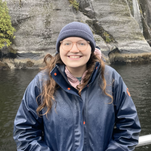 Smiling woman in a navy rain jacket and beanie stands by a railing with a waterfall cascading down a rocky, forested cliff behind her