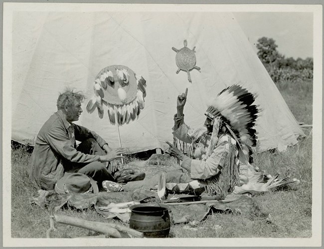An indigenous man in traditional dress raises a hand to speak using sign language. He is communicating with a non-indigenous man. Both sit on the ground in front of a tipi.