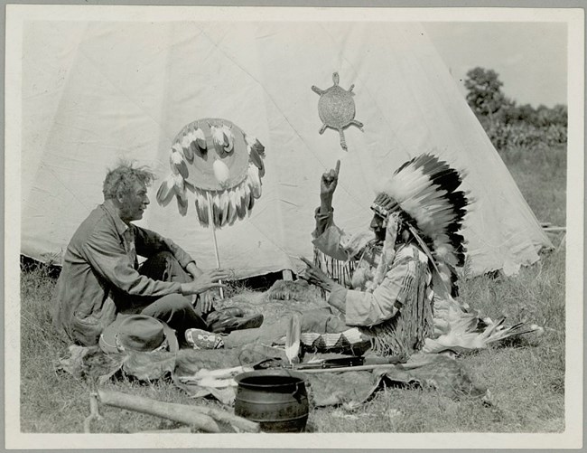 American Indian Sign Language (1) An indigenous man in traditional dress raises a hand to speak using sign language. He is communicating with a non-indigenous man. Both sit on the ground in front of a tipi.
