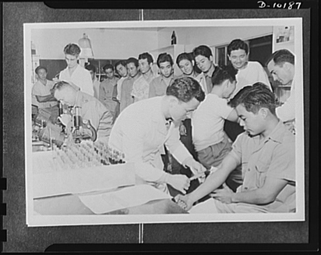 Black and white photo of a white man taking blood from the arm of a young Japanese American man. A microscope and vials of blood samples are arranged in a box on the table next to them. Other young Japanese American men wait in line behind them.