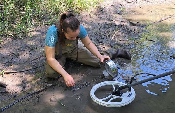 Woman wearing waders is holding an instrument for a mussel survey at Cub Creek. Park ranger is in the back standing next to the meadow and creek.