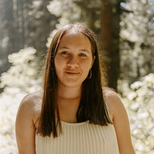 Woman with brown hair smiles in front of blurred redwood trees.