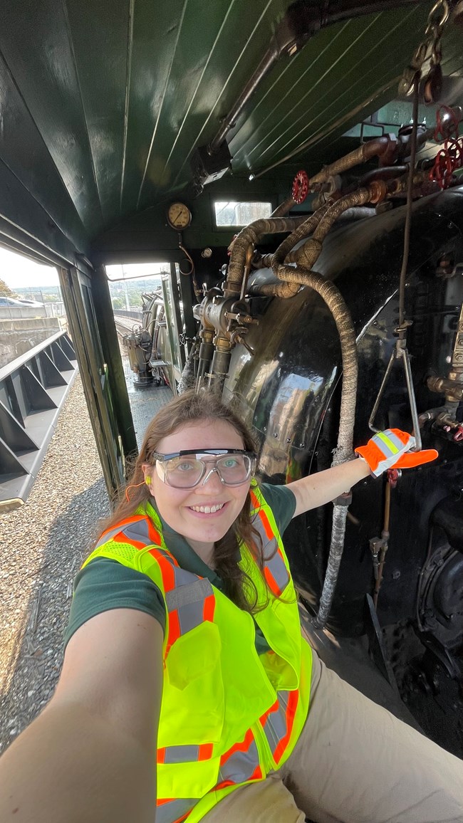 Smiling woman in safety goggles, a high-visibility vest, and gloves takes a selfie inside a locomotive cab, surrounded by pipes, gauges, and controls.