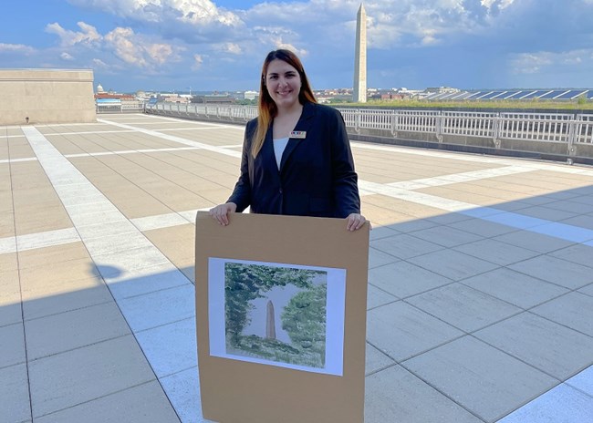 Amie Sardinha person holding a picture of a painting of the Washington Monument, with the Washington Monument in the background