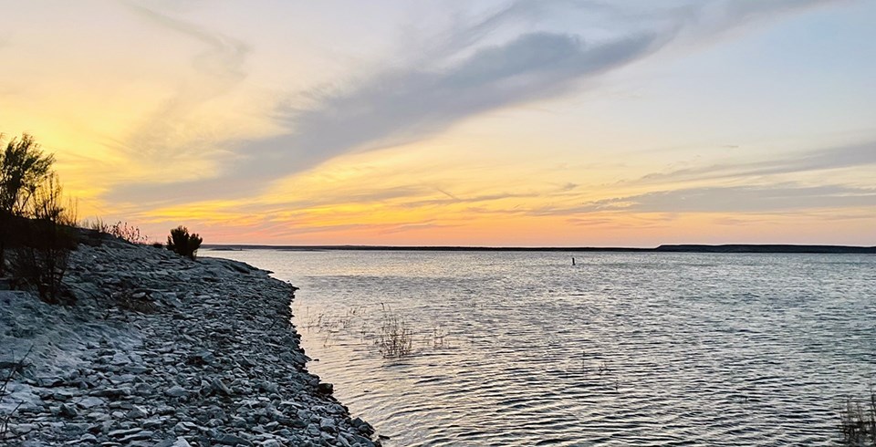 A rocky shoreline and a large rippling lake under an orange and yellow sky.