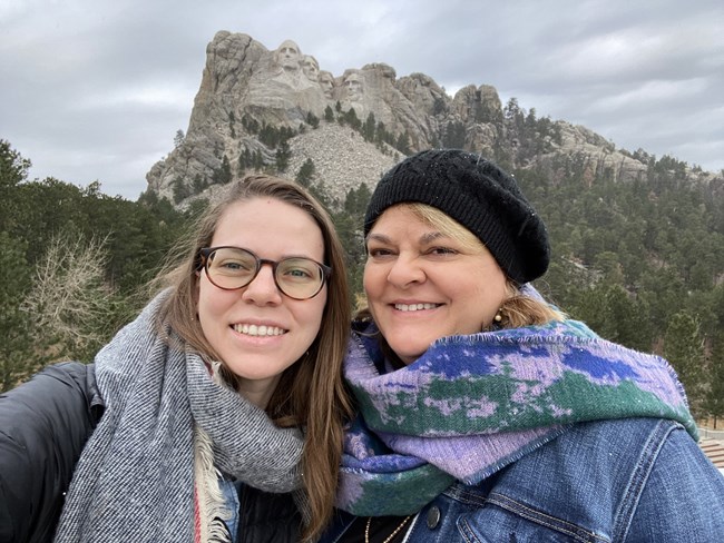 Amy and her mom posing for a photo in front of Mount Rushmore National Memorial on a road trip in 2021. Amy and her mom stand smiling wearing scarves in front of Mount Rushmore National Memorial on a cold, gray day.
