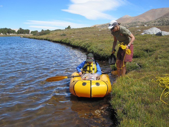 Andi Heard helps launch raft to collect mid-lake sample. One woman wearing a life vest sits in a small raft at the edge of a lake while another woman stands on shore holding rope that is used to haul raft back to shore if any problems arise.