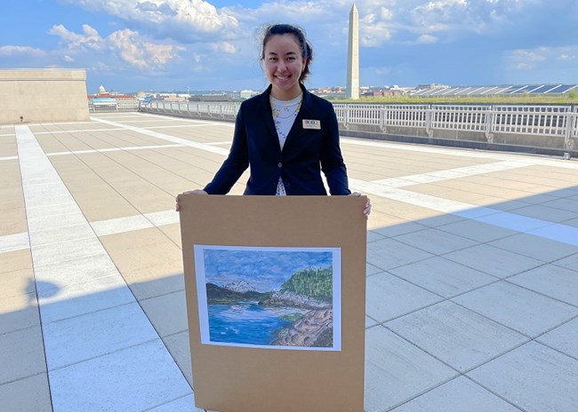 woman holding a painting of mountains and water, with Washington Monument in the background
