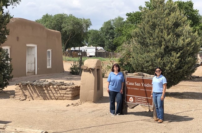 Two people stand next to a brown sign next to a historic adobe building.