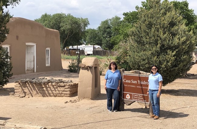 lt= Two people stand next to a brown sign next to a historic adobe building.
