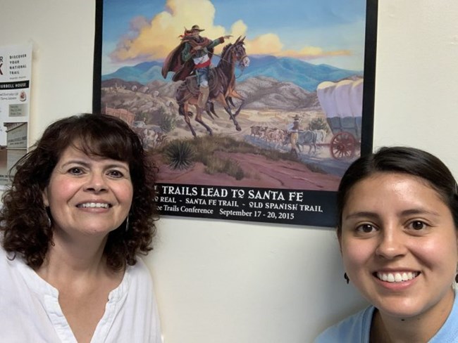 Two people stand next to a painting of a person on a horse.