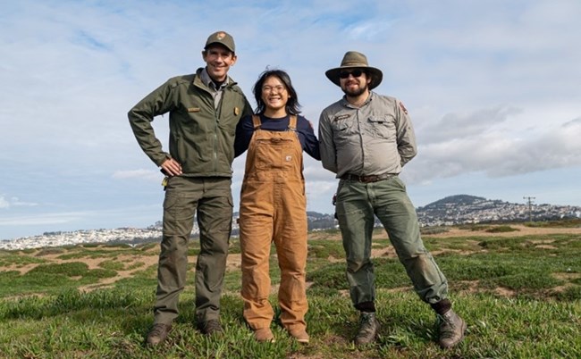 Three individuals outdoors in a grassy area wearing work clothes