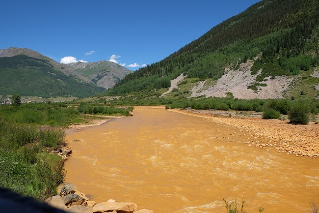 Animas River Mine Spill Landscape with tree covered mountains and river of orange water running through.