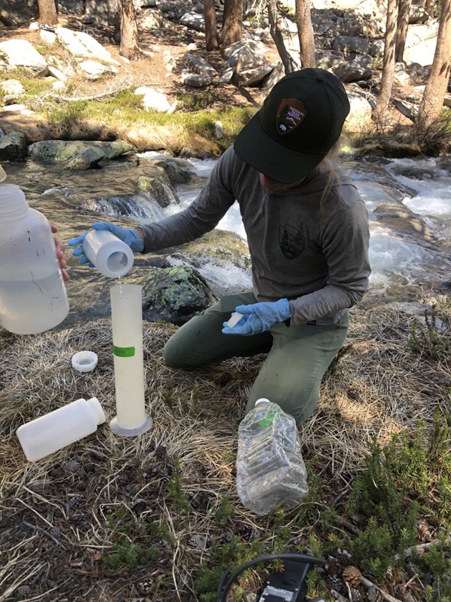 Scientist kneels at the edge of a river and pours liquid into a graduated cylinder, preparing for a salt dilution hydrology measurement.