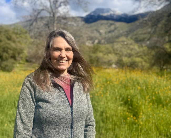 Smiling face of woman against a meadow and mountain backdrop.