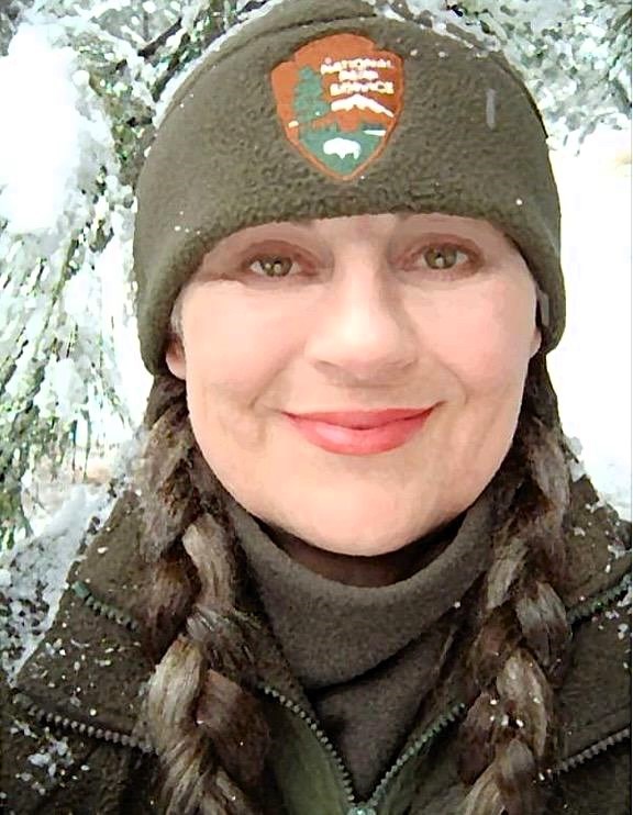Close up selfie of a smiling woman in a green, fuzzy NPS hat surrounded by snow.