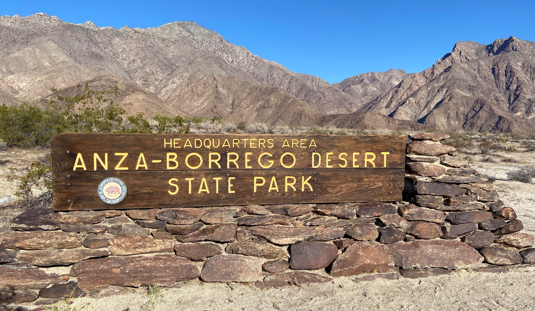 A rocky desert landscape with a wooden sign in the foreground and bare hills and ridges in the background. The sign reads Headquarters Area Anza-Borrego Desert State Park.