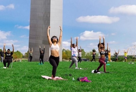 A group of primarily women in a yoga pose with arms stretched upwards on the green grass in front of the Arch's stainless steel leg in the background