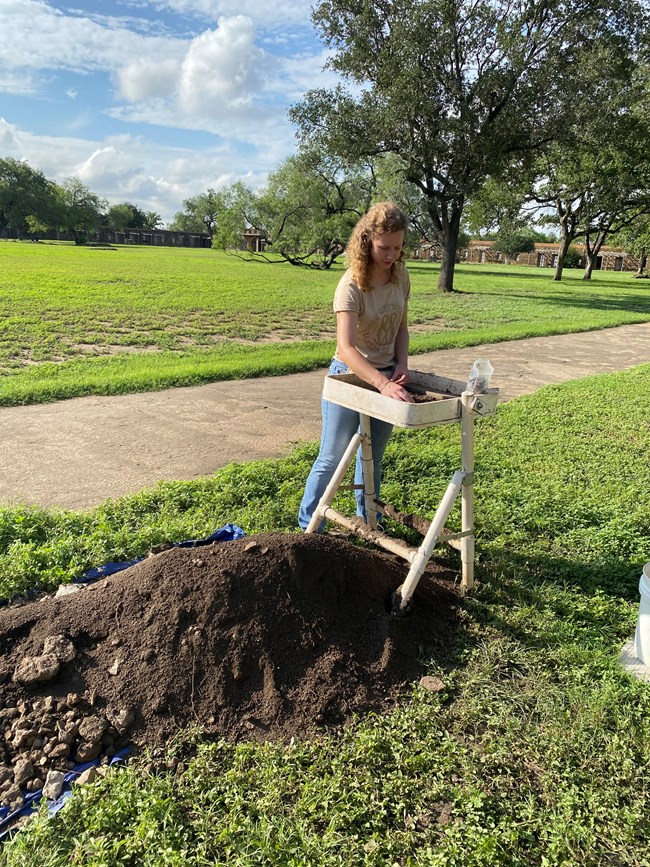 Woman stands with an archeology screen next to a pile of dirt.