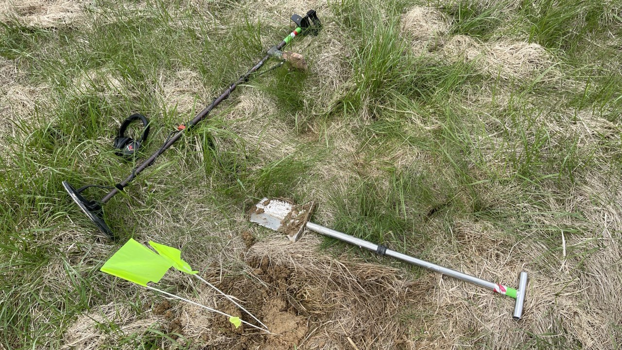 A metal detector, shovel, and yellow flags on grassy field.