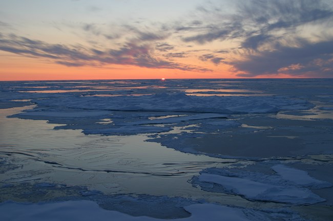 A setting sun casts an orange glow in the sky over an icy Arctic Ocean.
