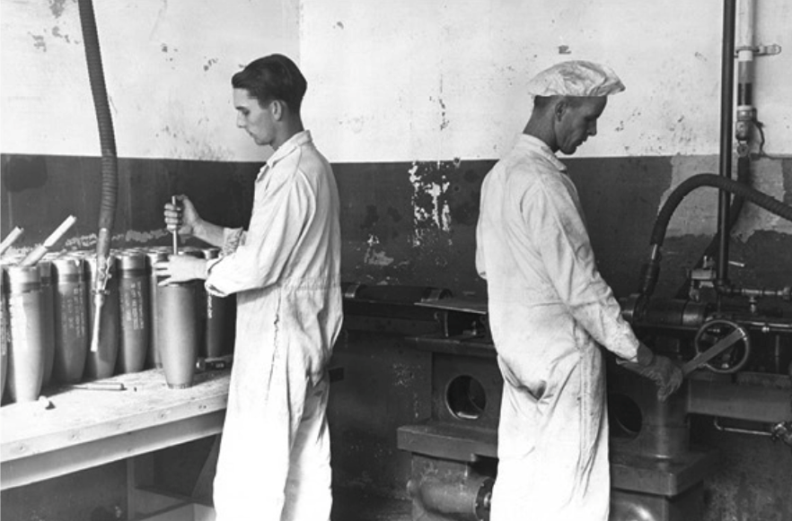 two men facing away from each other. Both are wearing white coveralls. One is wearing a shower-style cap. Both are working with large munitions shells.