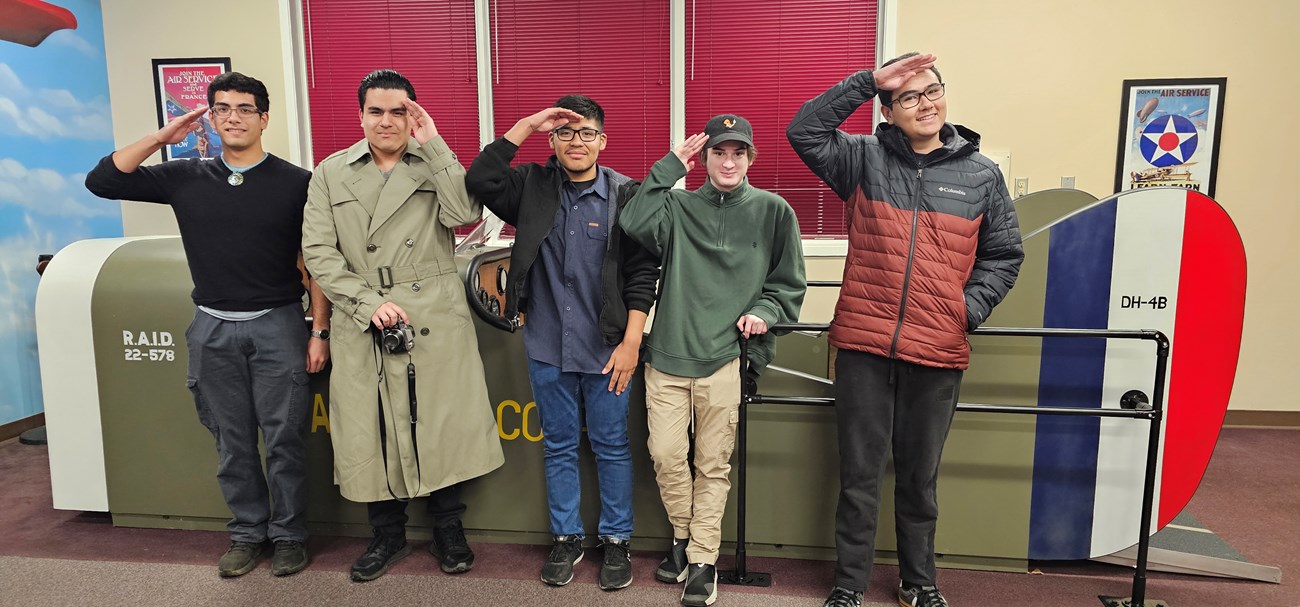 Five high school students stand in front of the aircraft model, saluting.