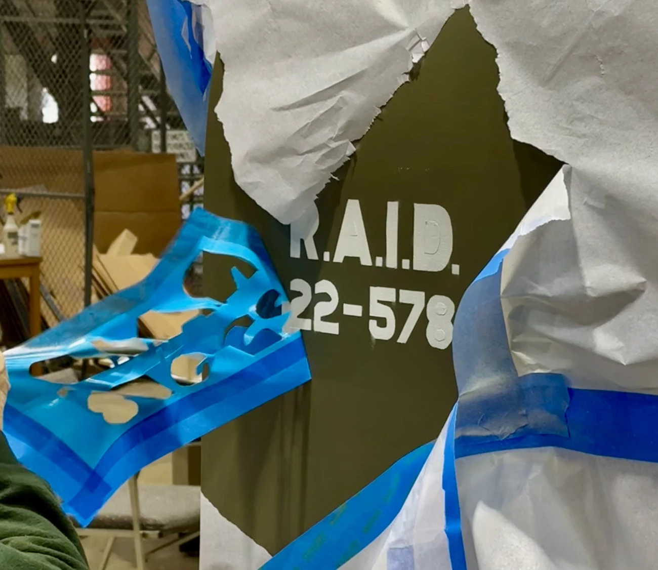 Liberty Plane stencil A close-up of a section of the airplane body shows a sticky, blue decal outline being pulled off after painting. White text reading R.A.I.D. 22-578 is on an olive green background.