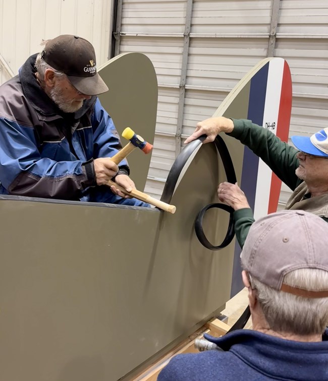 Three male volunteers apply black plastic edging to the wooden body of the aircraft model. Two are unspooling and guiding the roll of edging while one holds it in place with a wooden rod and uses a mallet to hammer it into place.
