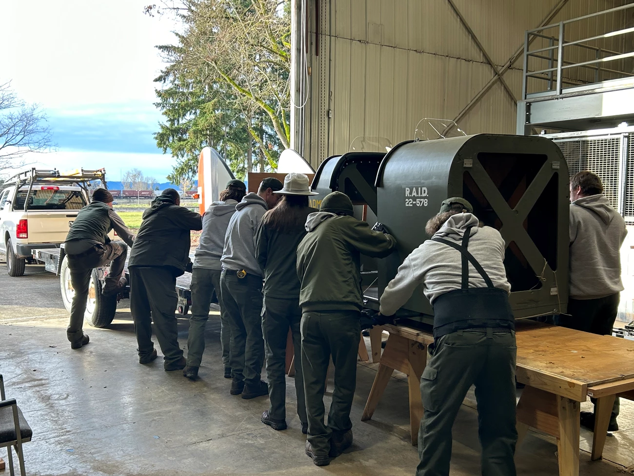 Liberty Plane move Twelve National Park Service staff, wearing uniforms, lift the aircraft from a wooden support onto a trailer for transport. They are in a warehouse with an open bay door.