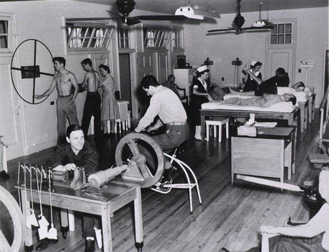 Nurses work with soldiers in therapeutic gymnasium. One patient uses stationary bicycle. Another soldier turns large wheel hung against wall. Two soldiers lay on raised tables with one leg elevated. One patient sits at table doing wrist exercises.