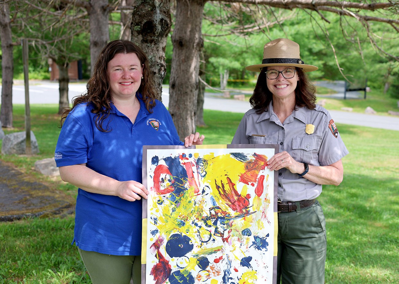 Two women, one in NPS uniform, hold up a colorful abstract painting in front of trees, grass and a road.
