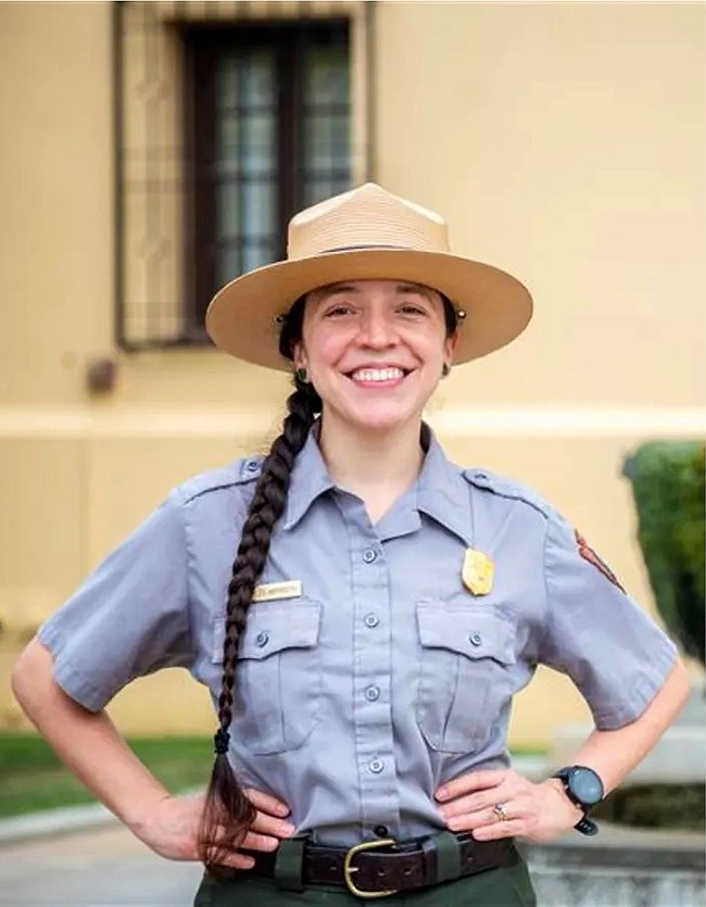 A smiling uniformed person, with dark hair in a braid, under a flat brimmed straw hat.