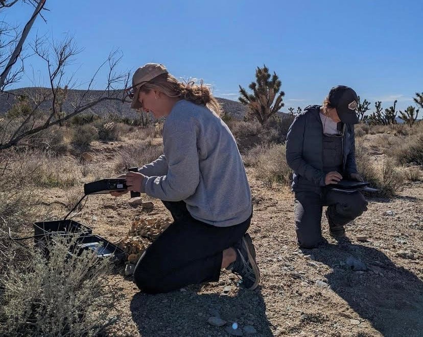 Two woman in desert landscape crouch down and hold electronic bat monitors, setting them up to record bat calls.