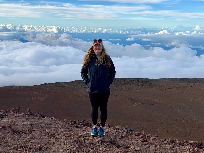 Woman stands on top of high peak, hands in pockets, with views of the tops of clouds and snowy mountain range behind her.