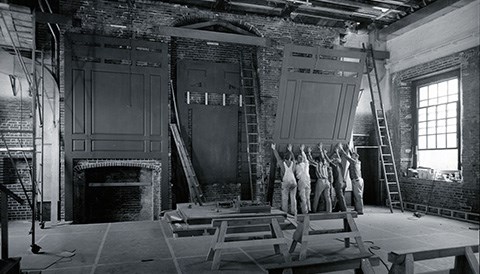 A Room paneling Four wood saw horses in a bare assemly room with five construction workers pushing a large piece of wood paneling in place on an interior brick wall.