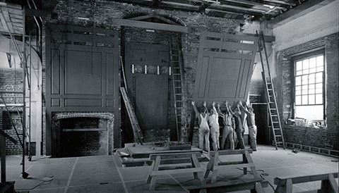 Four wood saw horses in a bare assemly room with five construction workers pushing a large piece of wood paneling in place on an interior brick wall.