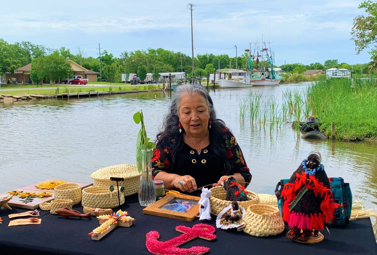 Atchafalaya National Heritage Area A woman weaving a basket sits at a table in front of a waterway. Table contains various objects representing regional culture including various baskets and dolls.