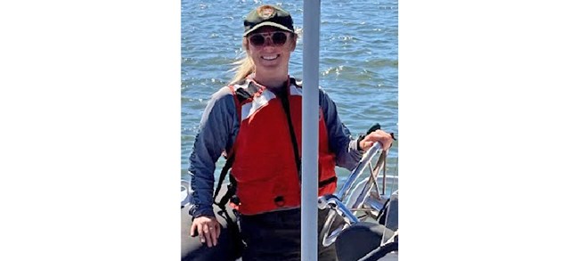 Kaitlyn Button Button in a NPS ball cap, orange life vest, and sunglasses, smiling at the helm of a boat.