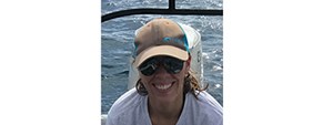 Smiling woman in sunglasses and ball cap and white shirt in a white boat with sunlit water behind her