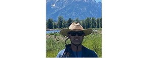 Man with large hat, blue shirt, and sunglasses  stands in sunlit meadow with snow-covered mountains in the background