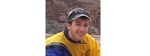 Smiling man in ball cap with yellow and blue jacket with brown water and hills behind him