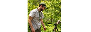 A man in a baseball hat  and grey t-shirt looks at a camera resting on a tripod. He is surrounded by green, leafy vegetation.