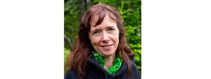 Woman with long red hair, wearing a blue jacket and a green shirts stands in front of a forest