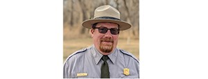 Man with red beard in NPS uniform stands in front of a field of dry grass