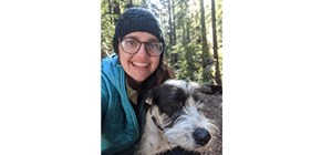 Janelle Christensen Woman in blue jacket with blue knit hat holds a black and wither terrier in front of a stand of evergreen trees
