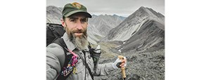 Man with beard and NPS uniform stands in front of a mountain range, holding a hiking stick