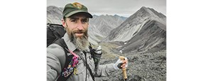 Josh Spice Man with beard and NPS uniform stands in front of a mountain range, holding a hiking stick