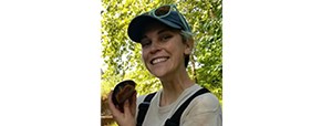 Smiling woman in waders and a ball cap holds up a turtle while standing in front of a stream flanked by green trees