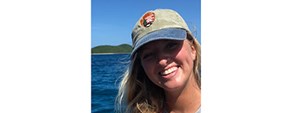 Kristen Ewen Smiling woman with blond hair blowing in the wind and an NPS ball cap in a boat with sunlit blue ocean and distant green shore behind her